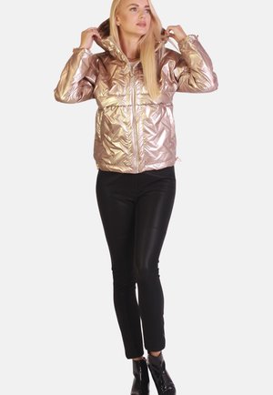 Frau trägt eine metallisch roségoldene Kapuzenpufferjacke, schwarze enganliegende Hosen und schwarze glänzende Stiefeletten und passt die Kapuze der Jacke an.