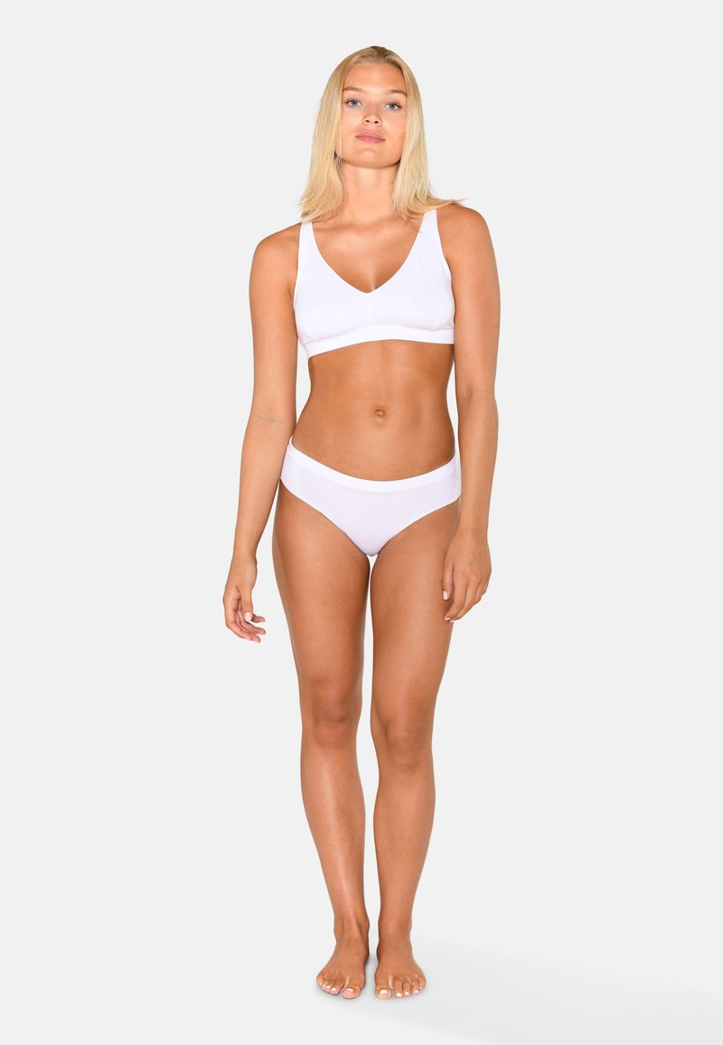 Femme aux cheveux blonds, debout pieds nus, portant un soutien-gorge de sport blanc et un sous-vêtement blanc sur un fond clair uni.