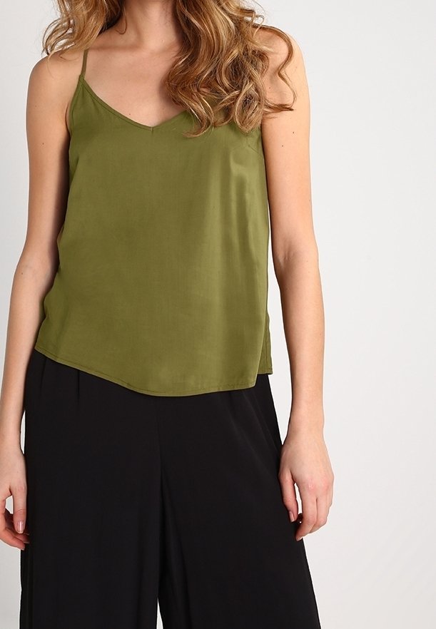 Femme portant un top sans manches vert olive à fines bretelles et un pantalon large noir, debout devant un fond uni.