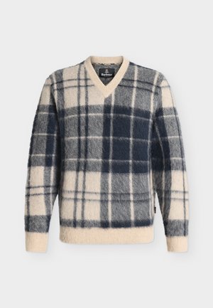 Jeune homme portant un pull à carreaux beige et noir, pantalon large beige, baskets blanches, tenant une veste beige dans une main, debout sur fond blanc.
