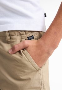 Pantalones cortos de algodón en color beige con un bolsillo trasero, un ligero brillo y costuras cosidas. Presenta una etiqueta azul marino con "Petzl Industries" en el bolsillo.