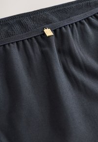 Gros plan sur un tissu noir avec une ceinture élastique froncée comportant un petit charme rectangulaire doré avec la lettre N gravée.