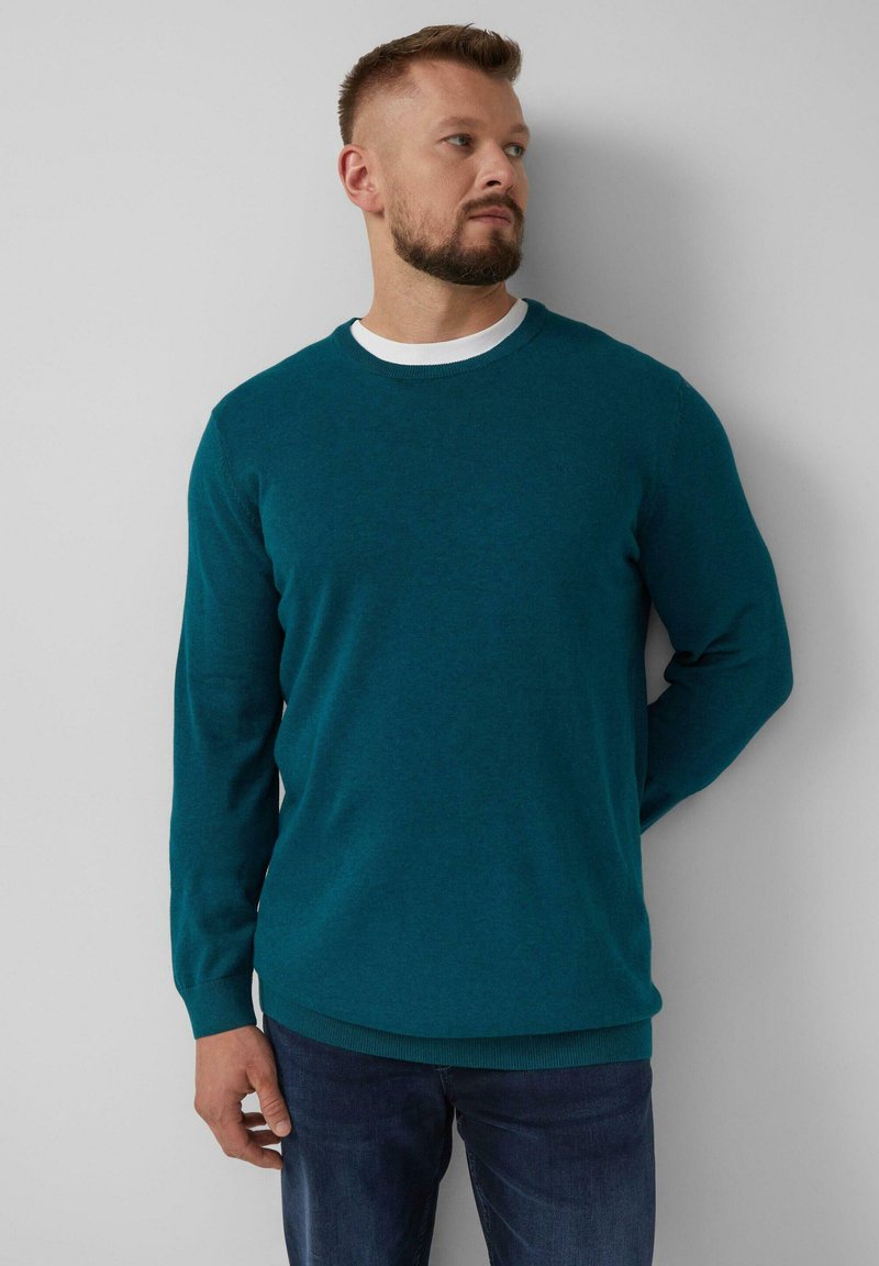 Türkiser Strickpullover mit Rundhalsausschnitt und langen Ärmeln. Der Stoff wirkt weich mit einer leicht strukturierten Oberfläche. Über einem weißen Hemd getragen.