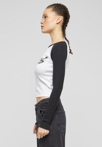 Bavlněný crop top s kontrastními černými dlouhými rukávy a bílým tělem. Na přední straně má grafický design a přiléhavý střih.