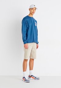 Langärmliges, marineblaues Hemd, beige Shorts, weiße Socken und Turnschuhe mit orangefarbenen und navyblauen Akzenten. Einfaches Design auf der Vorderseite des Hemdes.