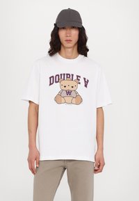 Camiseta blanca de algodón con un gráfico de un oso y "DOUBLE A" en púrpura encima. Corte holgado con mangas cortas. Se lleva con pantalones beige y gorra gris.