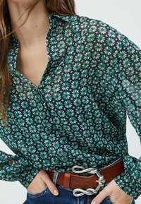 Genomskinlig svart blus med ett blommönster i grönt och vitt, med en knappkrage och långa ärmar, tillsammans med ett brunt bälte.