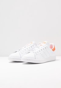 White leather sneakers with a smooth texture, featuring perforations for ventilation, a vibrant orange heel accent, and flat laces.
