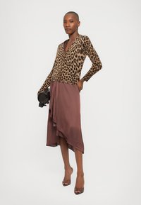 Leopard-print long-sleeve top in brown and black, paired with a brown asymmetrical silk dress, and black clutch bag, on a model with heels.