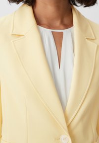 Blazer jaune clair avec des revers crantés, une fermeture à un bouton et un tissu lisse, associé à un haut blanc avec un décolleté en trou de serrure.