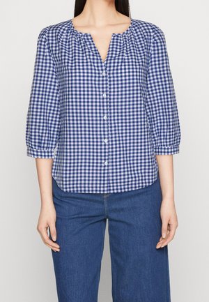 Frau trägt eine blau-weiß karierte Bluse mit Knöpfen und dreiviertellangen Ärmeln, kombiniert mit blauen Jeanshosen.