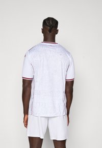 Weißes Fußballtrikot mit kurzen Ärmeln und roten Akzenten, mit einem strukturierten grafischen Design auf dem Rücken. Einfache Shorts in passendem Weiß.