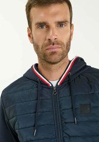 Chaqueta de abrigo con capucha en color navy, diseño acolchado, detalles en franjas rojas y blancas en el cuello, y cremallera plateada; incluye un parche de logo en el pecho.