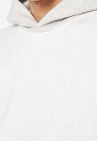 Gros plan d'une personne portant un sweat à capuche gris clair avec un logo Jumpman blanc brodé discrètement sur la poitrine.