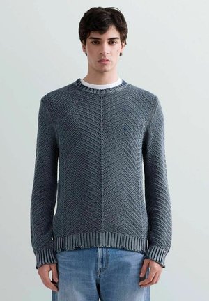 Giovane uomo che indossa un maglione a maglia strutturata blu sopra una camicia bianca, abbinato a jeans azzurri, in piedi davanti a uno sfondo semplice.