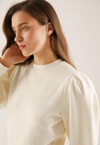 Jonge vrouw met lang bruin haar die een crème kleurige top met opgeblazen mouwen draagt, zij kijkt opzij met haar hand vlakbij haar oor.