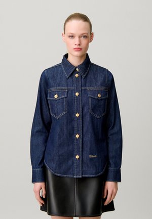 Femme portant une chemise en jean bleu foncé à boutons dorés et une jupe en cuir noire sur un fond gris uni.