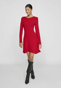 Sarkans ribots adītas kleitas ar garajām piedurknēm, izplūdušu apakšmalu un zelta pogu akcentiem uz pleciem. Saskaņota ar augstiem melniem ādas zābakiem.