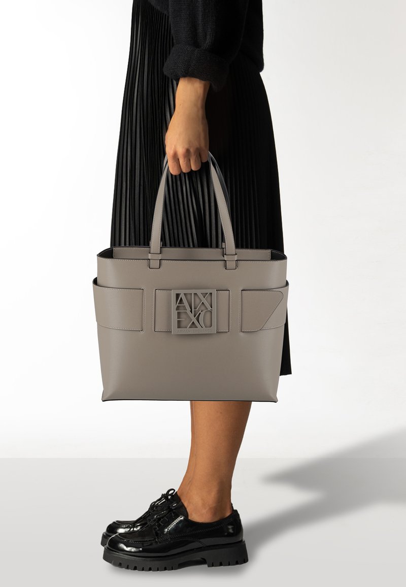 Borsa a mano strutturata grigia con due manici superiori, grande tasca anteriore e dettaglio del logo. Materiale liscio e accenti geometrici arricchiscono il design.