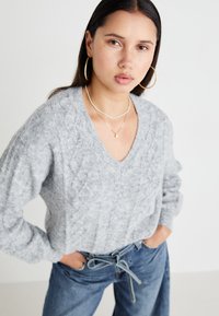 Pull en laine gris avec un col en V, motifs texturés et manches longues. Porté avec un jean taille haute bleu avec une taille nouée.