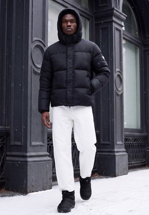 Jeune homme portant une doudoune noire avec capuche, un pantalon blanc et des bottes d’hiver noires, marchant sur un trottoir enneigé à côté d’un bâtiment sombre.
