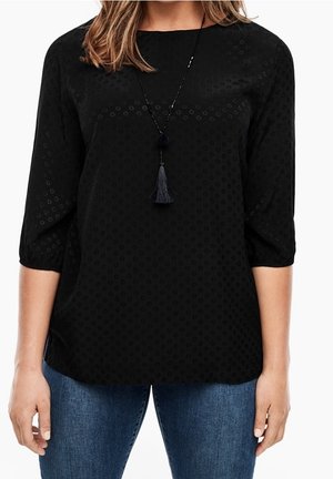 Mujer con camiseta negra texturizada de manga tres cuartos y un collar negro con borlas, y pantalones vaqueros azules, de pie con los brazos relajados a los lados.