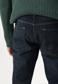 Jeans in denim blu scuro con cuciture dorate, caratterizzati da un classico design a cinque tasche e una leggera texture. Un maglione verde a coste copre parzialmente l'elastico in vita.