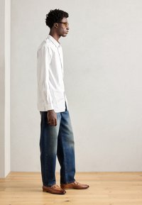Cămașă albă cu dungi, cu mânecă lungă, blugi din denim albastru, ușor decolorati, și mocasini din piele maro cu accente argintii. Modelul este poziționat pe un fundal neutru.