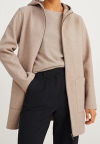 Abrigo de mezcla de lana color beige con capucha, cremallera frontal y bolsillo lateral. Lleva un top beige a juego y pantalones negros. Diseño simple.