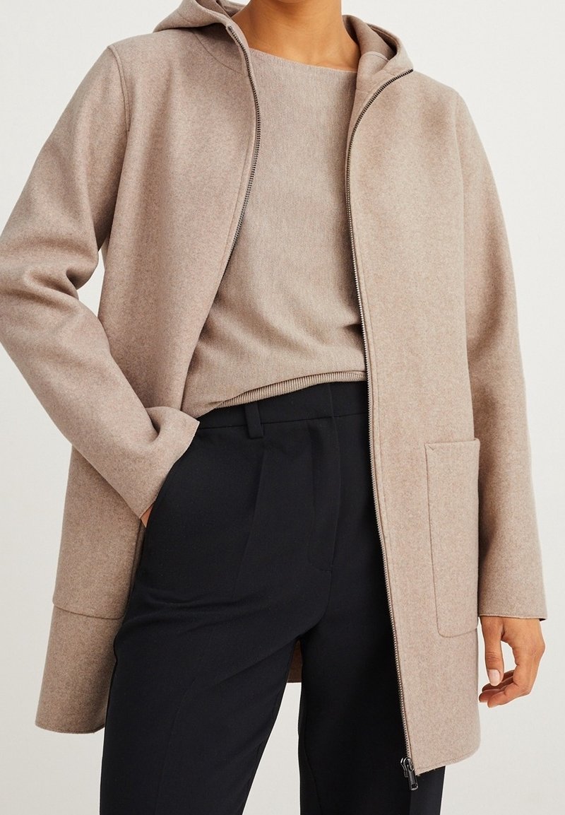 Abrigo de mezcla de lana color beige con capucha, cremallera frontal y bolsillo lateral. Lleva un top beige a juego y pantalones negros. Diseño simple.