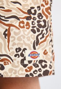Tessuto beige con motivo leopardato in tonalità di marrone e marrone scuro. Include un'etichetta bianca cucita con il logo "Dickies".