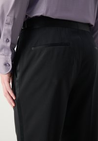 Black tailored trousers with a smooth texture, featuring a sleek waistband, subtle side stripe, and a single back pocket.
