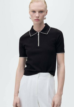 Zwarte polo met korte mouwen, witte bies en ritskraag; gedragen met witte broek. De stof lijkt glad en aansluitend.