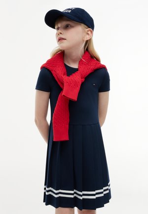Fille portant une robe bleu marine avec des rayures blanches près de l'ourlet, un pull rouge drapé sur ses épaules et une casquette de baseball bleu marine, regardant vers la gauche.