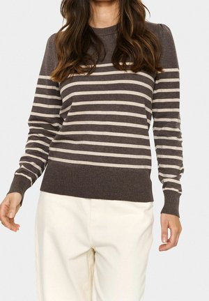 Kvinde iført en brun og beige trøje med horisontale striber sammen med off-white bukser, vist fra skuldre til midt på låret.