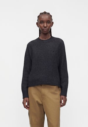 Ung kvinde med flettet hår iført en mørkegrå sweater og lysebrune bukser, stående mod en ensfarvet hvid baggrund.