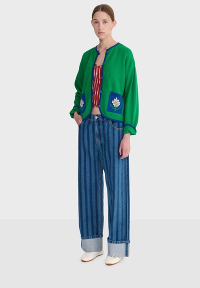Femme portant un cardigan vert avec des bordures bleues et des poches à fleurs, un haut rayé rouge, un jean large à rayures bleues avec revers, et des chaussures blanches, debout.