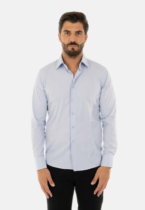 Uomo con la barba che indossa una camicia azzurro chiaro abbottonata e pantaloni neri, in piedi dritto davanti a uno sfondo bianco uniforme.