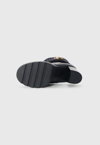 Black heeled shoe with a ribbed textile upper, gold accent details, and a textured rubber sole featuring horizontal grooves for traction.