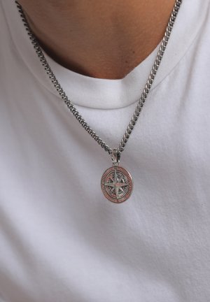 Sudraba ķēdes kaklarota ar apaļu kompass piekariņu, kuram ir gravēti detaļas un sarkans akcents, valkāta virs baltas krekla.