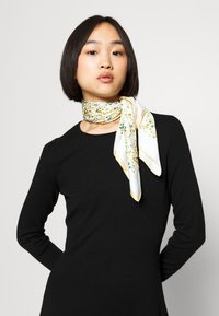 Cachecol de seda em branco com um padrão floral apresentando acentos em amarelo e verde. Com uma borda dourada fina, atado ao pescoço.