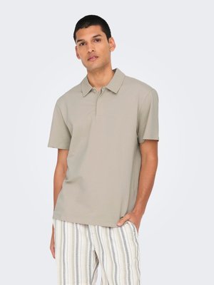 Mand iført en lysebeige kortærmet polo og beige, hvide og grå bukser med lodrette striber, stående med den ene hånd i lommen.