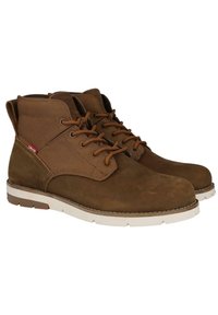 Botines de ante marrón con un cuello alto, cierre de cordones en la parte delantera y suela de goma blanca. Con una etiqueta de logo en el costado y detalles de costura.