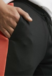 Black athletic pants with a textured fabric, featuring a contrasting orange panel. Hand inserted into pocket for detail.
