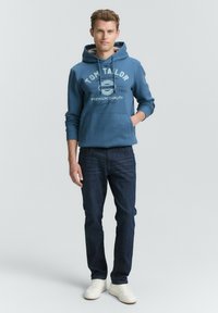 Blaue Kapuzenjacke mit Aufdruck, kombiniert mit dunkelblauen Jeans. Das Modell trägt weiße Sneakers und zeigt einen lässigen Stil.