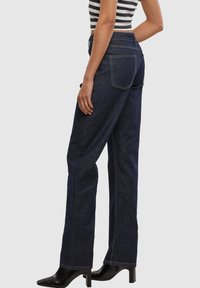 Frau trägt hoch taillierte dunkelblaue Jeans mit weitem Bein, gestreiftes ärmelloses Oberteil und schwarze Stiefeletten mit Absatz, steht vor einem schlichten Hintergrund.