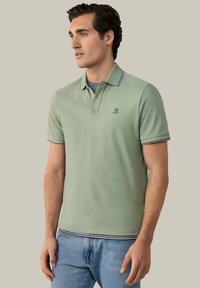 Homme portant un polo vert clair par-dessus un sous-pull rayé et un jean bleu, debout devant un fond neutre uni, regardant vers la gauche.