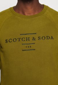 Olivengrøn sweatshirt med ribbede manchetter og halsudskæring. Sort printet tekst "SCOTCH & SODA 105" i en fed skrift på forsiden.