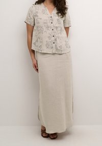 Světle béžová lněná košile na knoflíky s abstraktními liniemi, spárovaná s dlouhou sukní ve stejném odstínu. Hnědé sandály s pásky dotvářejí celý outfit.