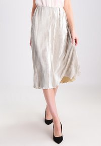 Jupe midi plissée en argent avec un ourlet légèrement évasé, dotée d'un tissu métallique texturé. Associée à des escarpins noirs.